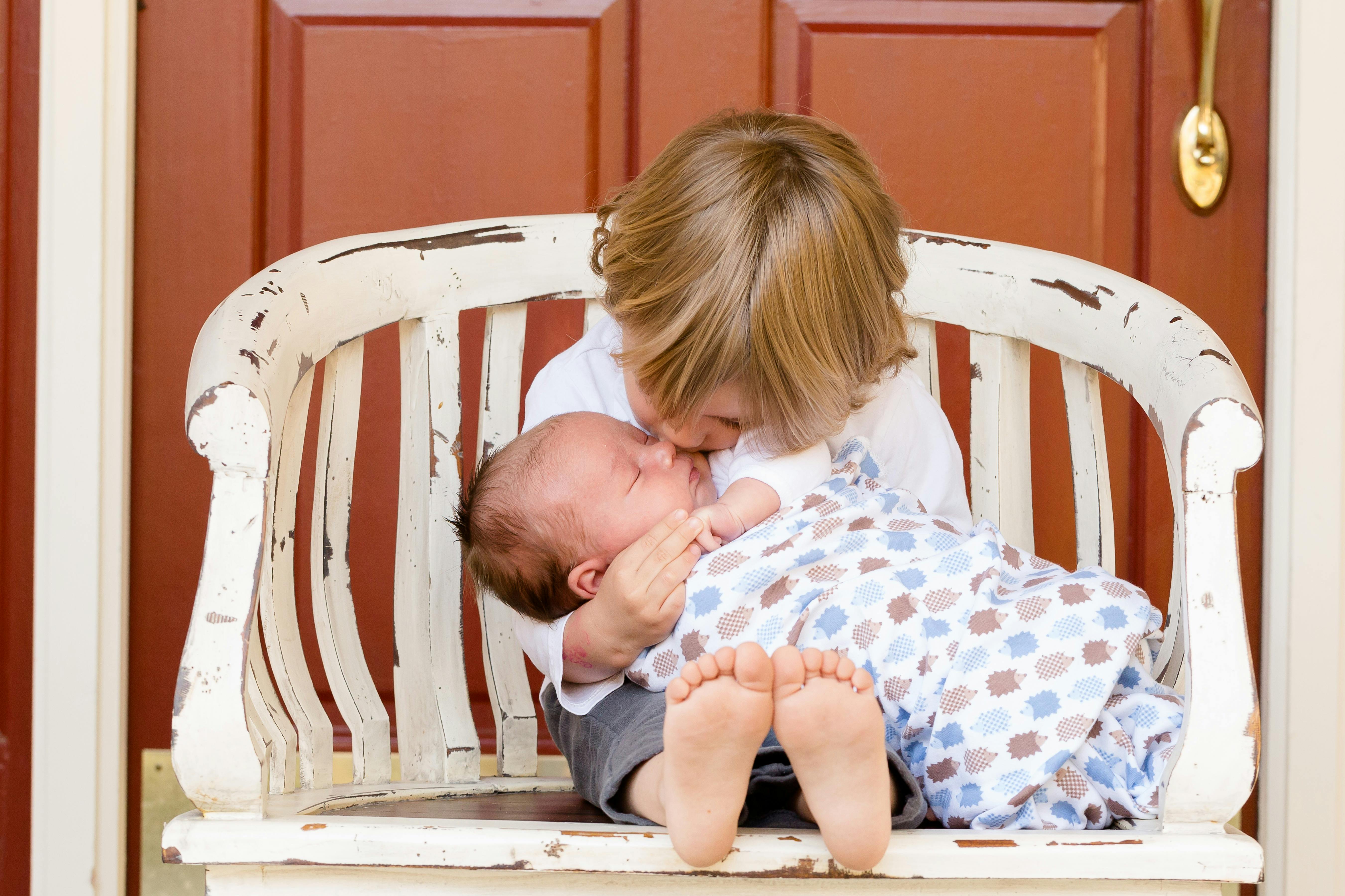 Toddler siting in a chair holding a baby and kissing its cheek