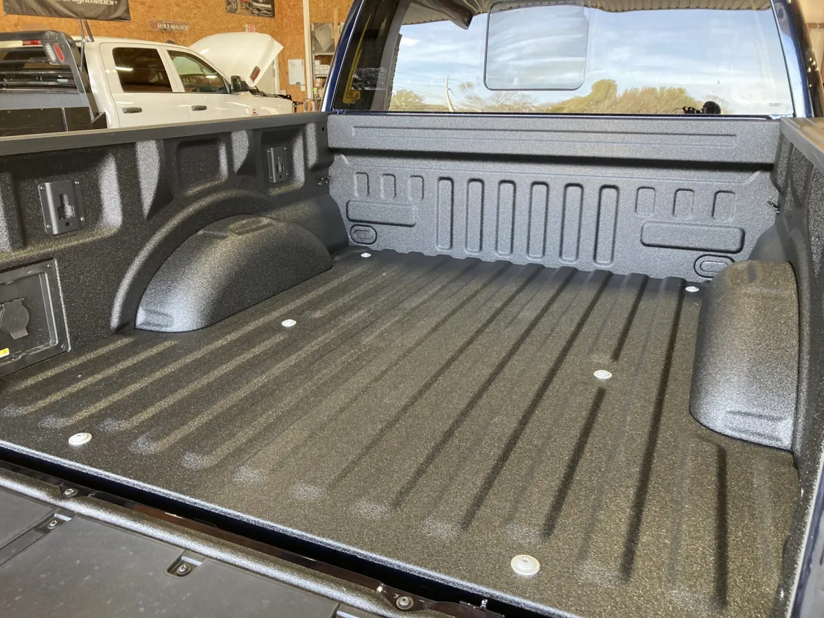 Interior of a pickup truck bed coated in a black textured spray‑on liner, showing wheel wells, tie‑down anchors, and corrugated floor ribs, with another truck and garage walls visible in the background.