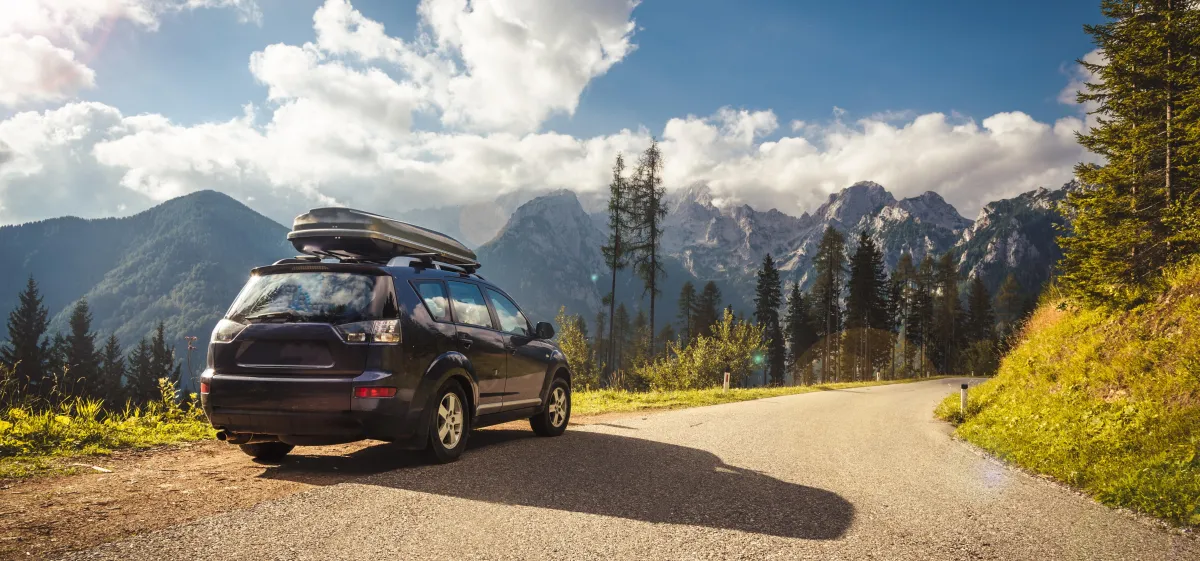 Dark SUV with a rooftop cargo box parked beside a winding mountain road, backed by forested peaks under a partly cloudy sky.