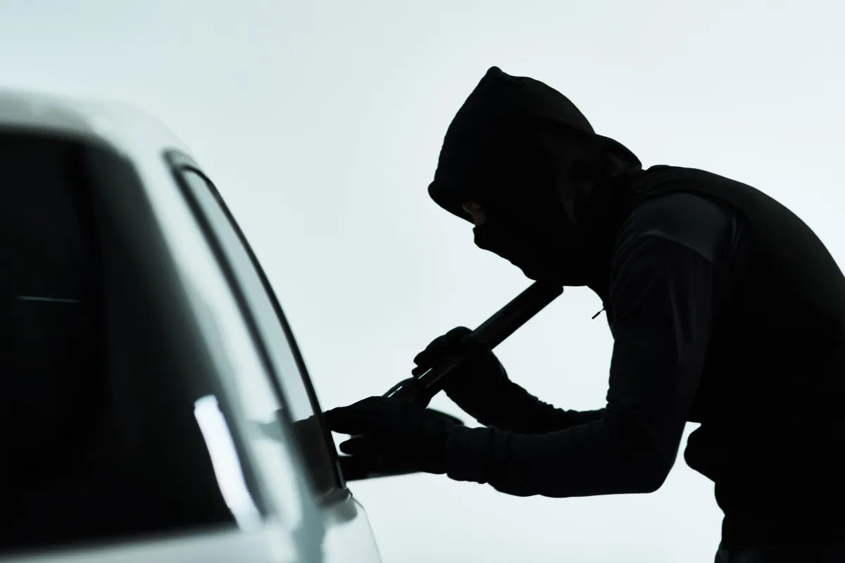 Silhouette of a masked person in a hood and gloves prying open a car window with a flashlight