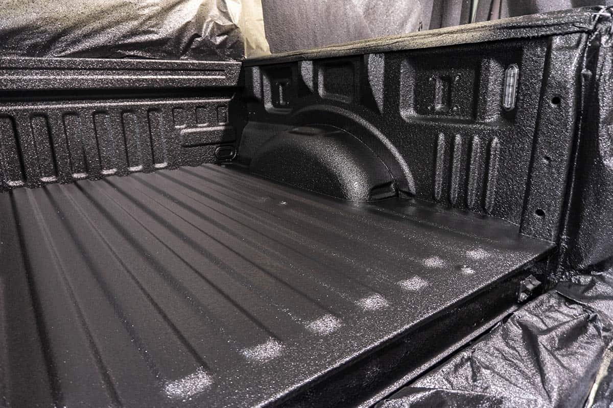 Interior of a pickup truck bed coated in a black textured spray‑on liner, showing wheel wells, tie‑down anchors, and corrugated floor ribs, with another truck and garage walls visible in the background.