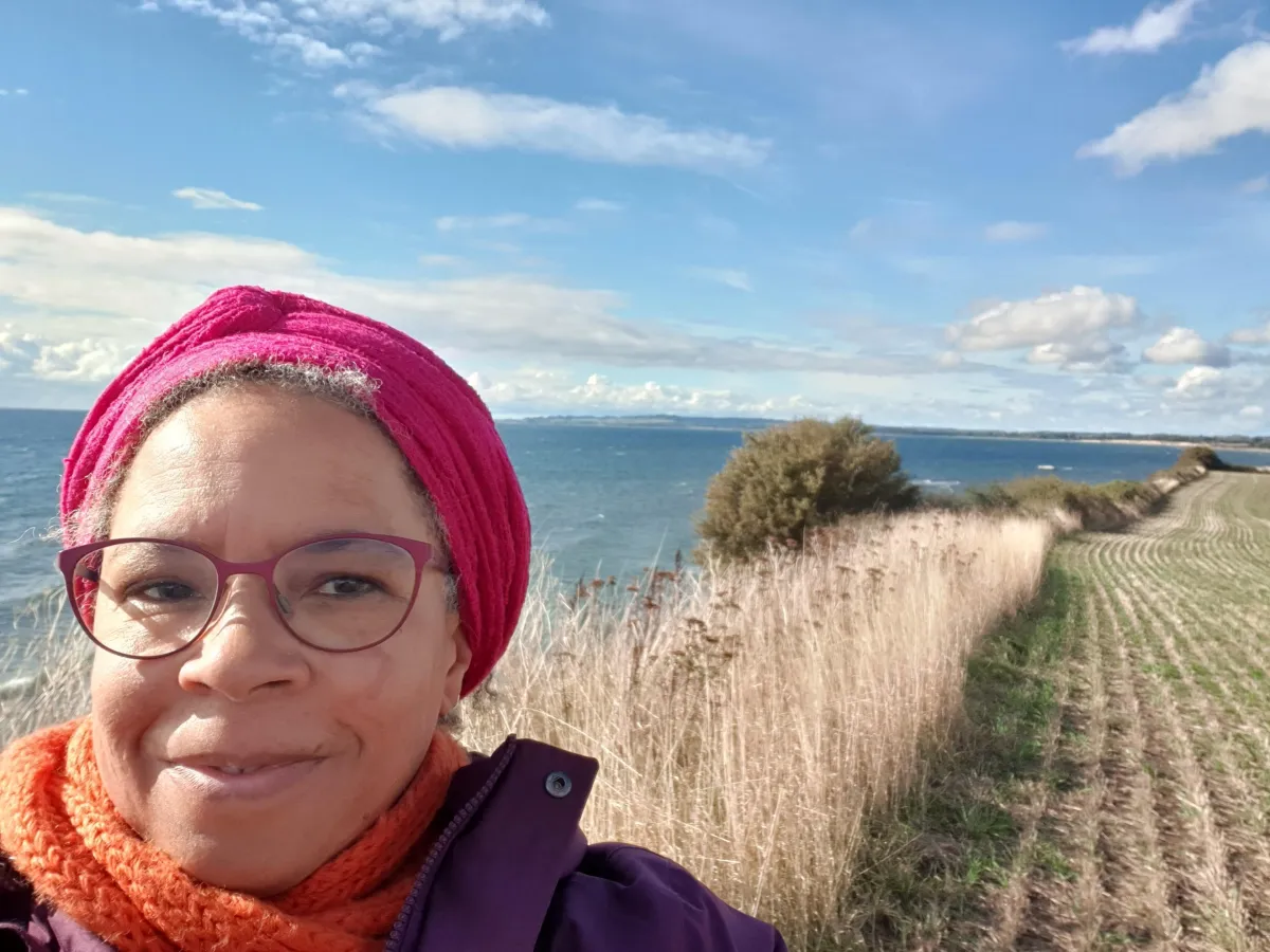 Tanya Spencer smiling in a windswept coastal field with the ocean behind her, wearing a pink headwrap, orange scarf, and purple jacket. Captures her vibrant, grounded leadership presence during a walk in coastal Denmark.