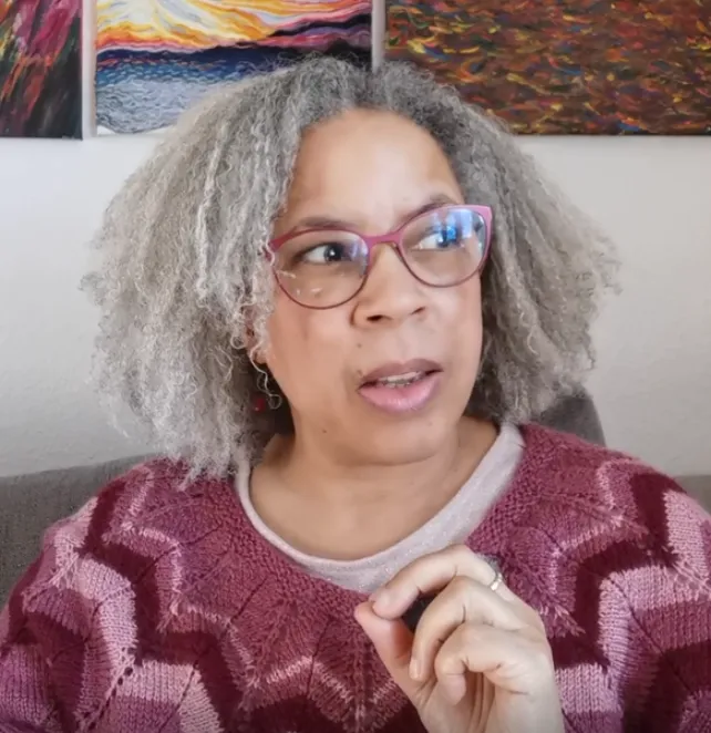 Tanya Spencer seated indoors, mid-sentence with a thoughtful expression and hand raised, wearing glasses and a maroon sweater. Her paintings hang behind her. Captures her expressive, strategic energy as a leadership communication expert and coach.