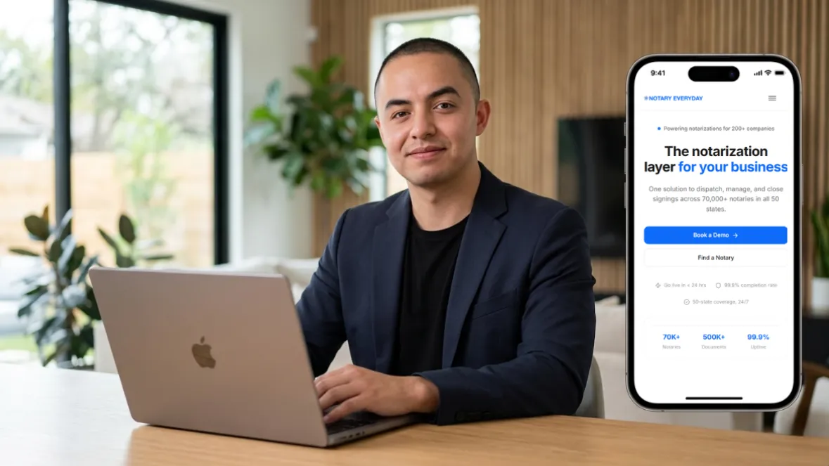 Man in his late 20s sitting at a clean desk with a laptop open, looking at the camera with a slight smile, with a blurred modern home office in the background.