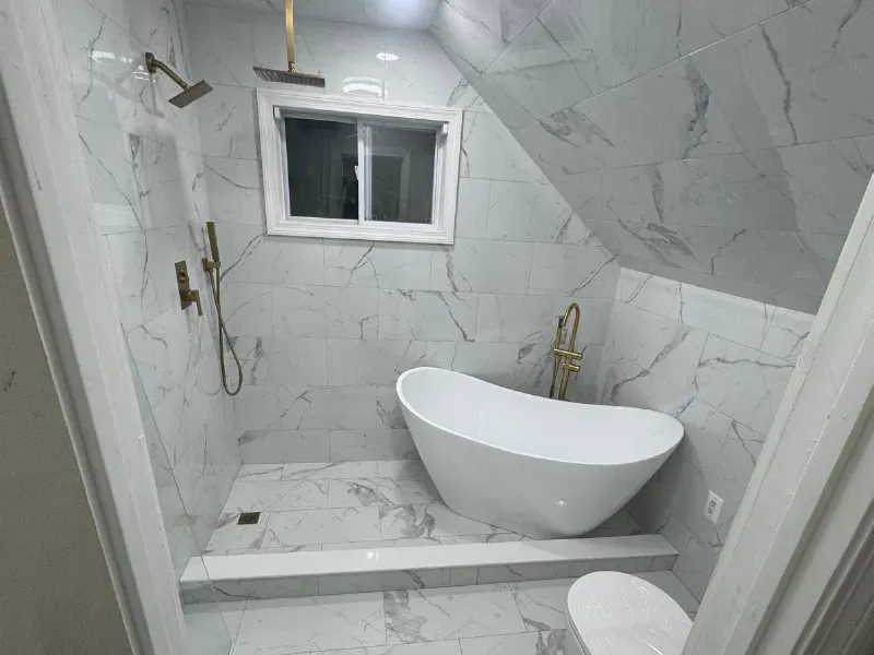 White marble tile covering walls, floor and sloped ceiling in luxury bathroom with freestanding tub and gold fixtures by CLA New England Tile – Greater Boston Area