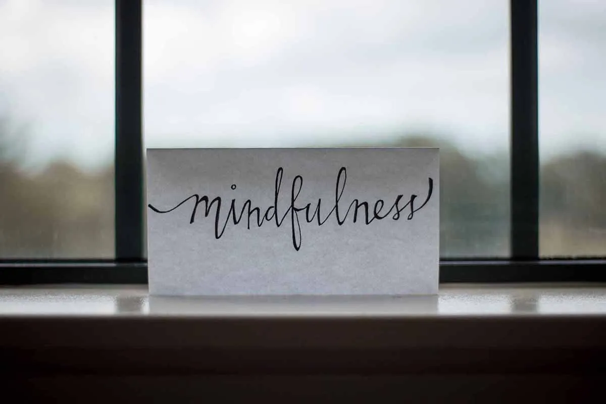A close-up shot of a small decorative card with the word "mindfulness" written in elegant script font, placed near a window sill to represent mental calm and holistic stress management.