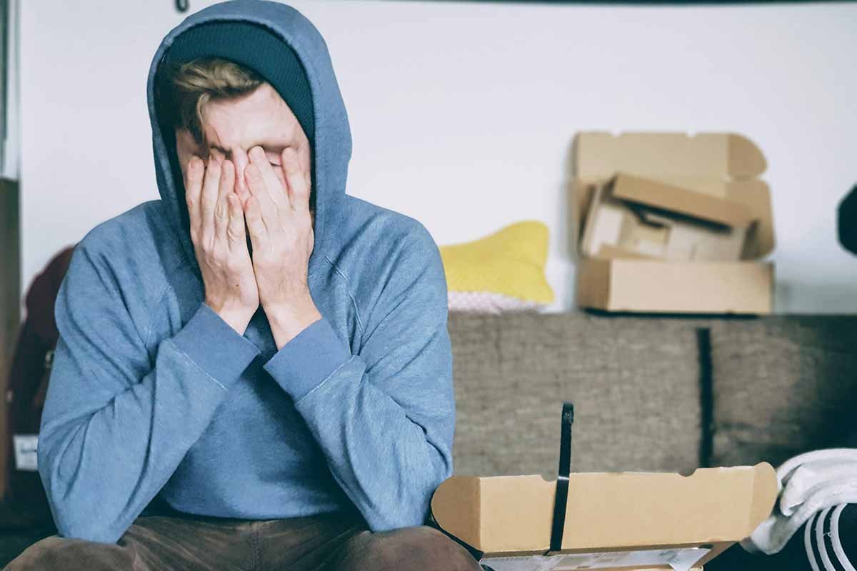 A tired young man sitting on a couch covering his face with his hands, representing chronic fatigue, stress, and low energy levels.
