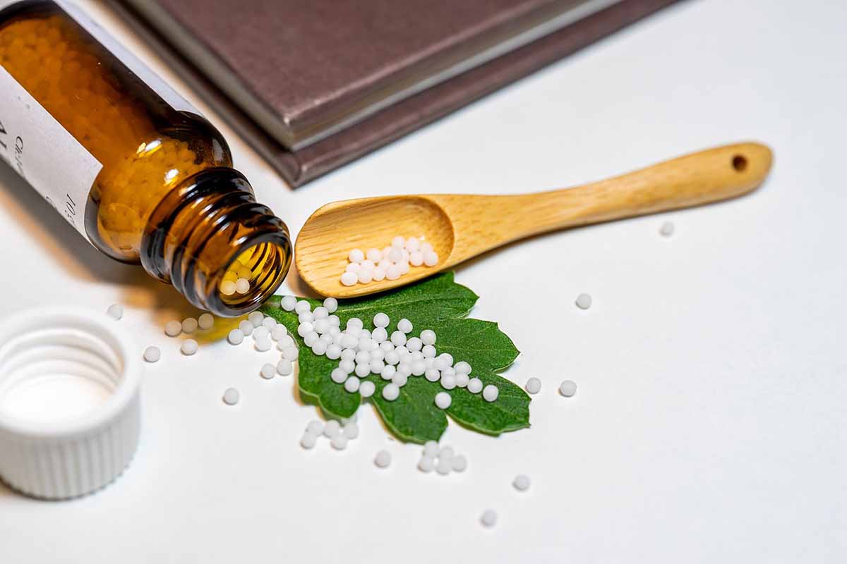 An amber glass bottle spilling tiny white homeopathic pellets onto a green leaf and a white surface next to a small wooden spoon, representing natural remedies and naturopathic treatments.