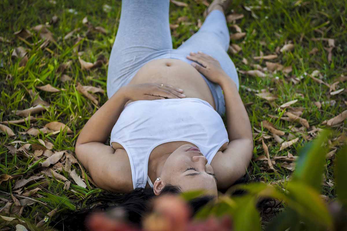 An overhead view of a happy pregnant woman lying peacefully on green grass, gently resting her hands on her baby bump to represent holistic prenatal care and natural fertility solutions.