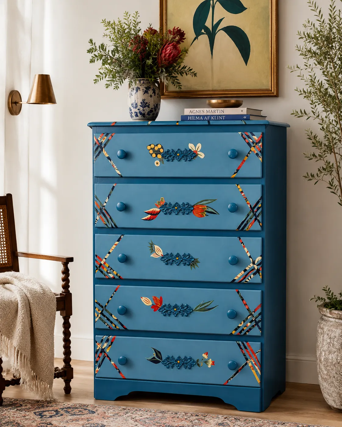A hand-painted blue floral dresser styled in a sophisticated European-inspired bedroom with pale ivory paneled walls, herringbone wood flooring, sheer window drapes, brass accents, a framed architectural artwork, and crisp white bedding with soft blue details. The dresser features colorful floral embellishments, patterned diagonal striping, blue knobs, and minimal elegant decor on top, including a cream vase with greenery and a small gold dish