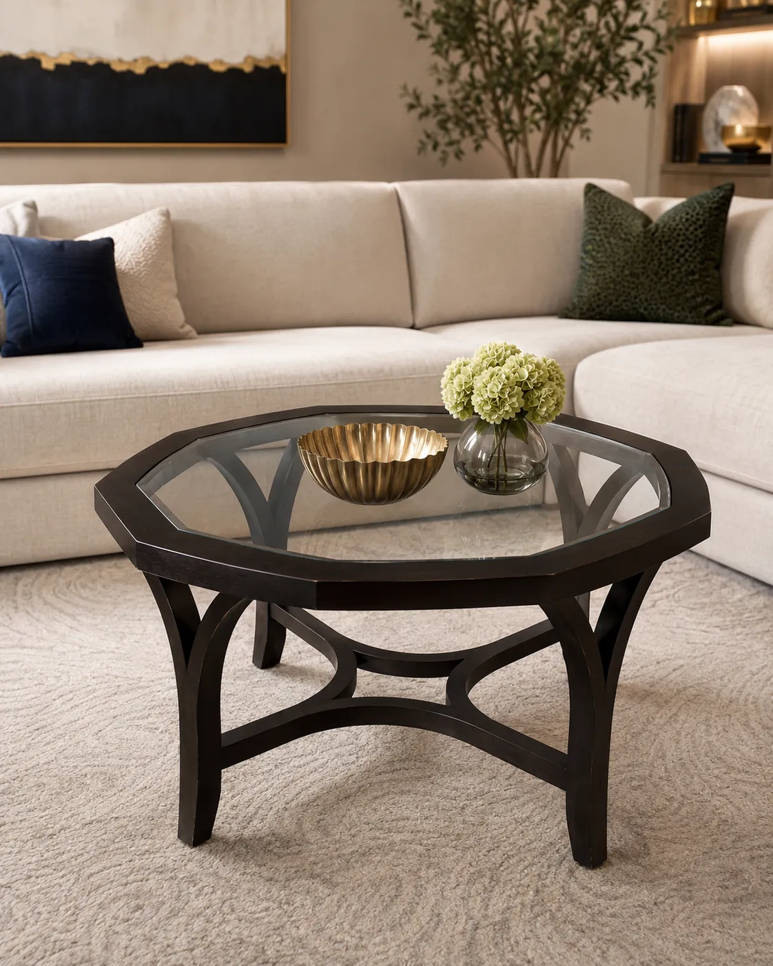 **Alt Text:** Dark real wood octagonal coffee table with a clear glass top, curved sculptural base, and elegant arched legs styled in a modern luxury living room. The table is centered on a soft neutral rug in front of a cream sectional sofa, with minimal decor including a gold accent bowl and a small glass vase of pale green flowers, highlighting the table’s clean lines, glass surface, and refined statement design.
