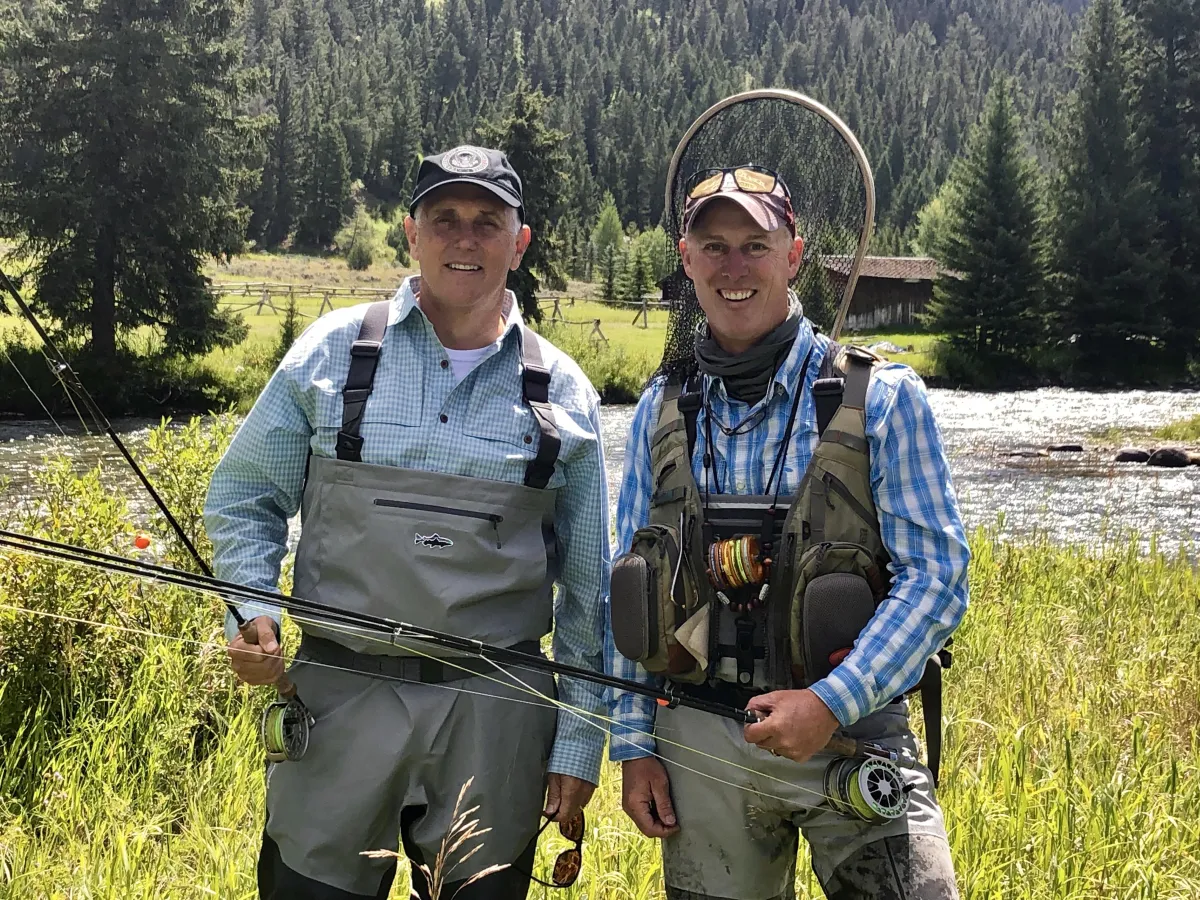 Darrel DeLeon and Vice President Mike Pence standing on a Montana riverbank, each holding a fly rod with forested hills in the background.