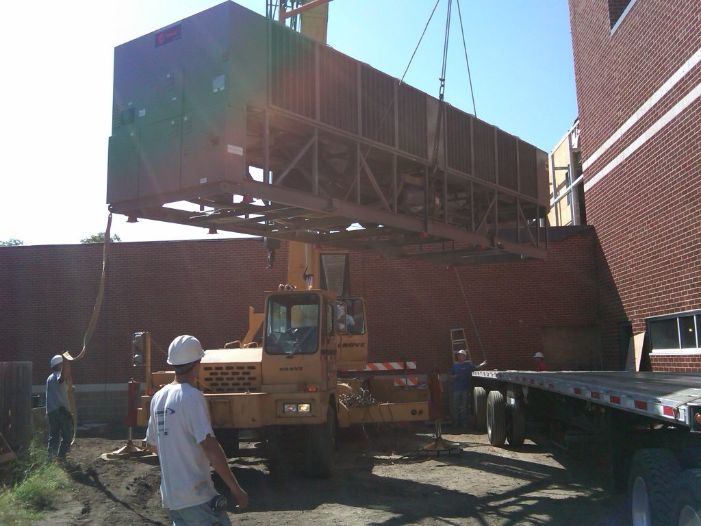 Crew lifting and installing a large commercial HVAC unit at an industrial building site using heavy equipment.