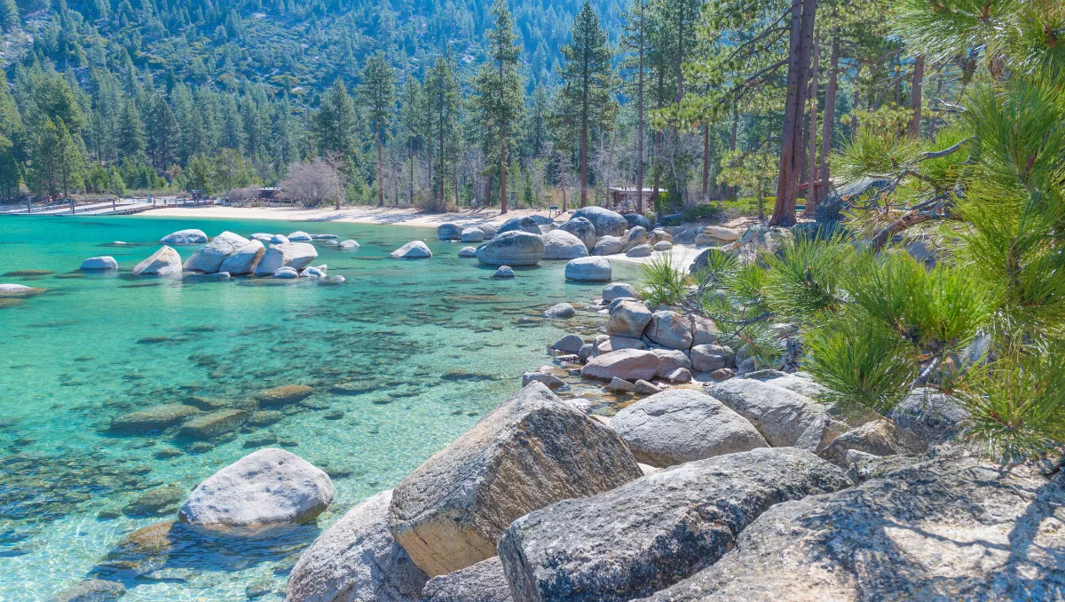 Lake Tahoe with crystal-clear turquoise water, rocky shoreline, pine trees, and snow-capped Sierra Nevada mountains under a blue sky.