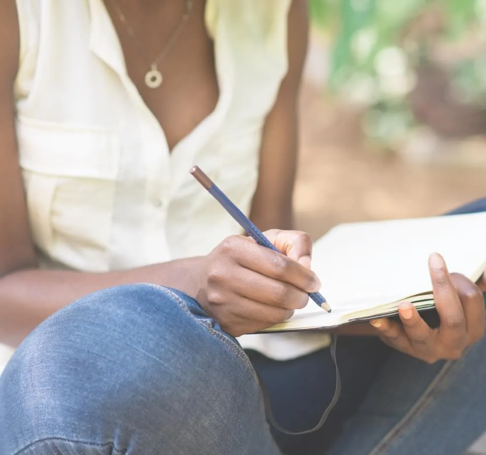 dark skinned woman's torso sitting crosslegged with a notebook and pencil in her lap