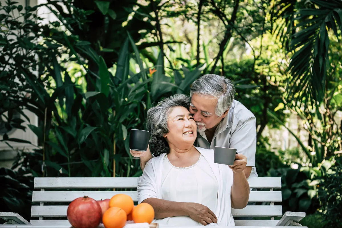 An older couple sharing coffee in a garden, with fresh fruit on the bench
