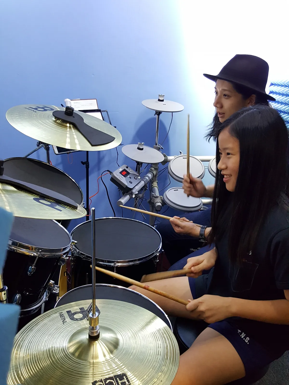 Smiling student seated at a drum kit holding drumsticks confidently in a modern music school studio
