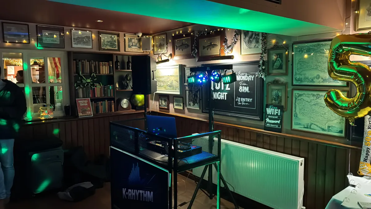 DJ booth setup at dusk with dual speakers, laptop showing waveform, LED uplights and fog haze blurred in background, wide 3:2 composition, photorealistic style demonstrating scale, safety rigging, and festival-grade production.