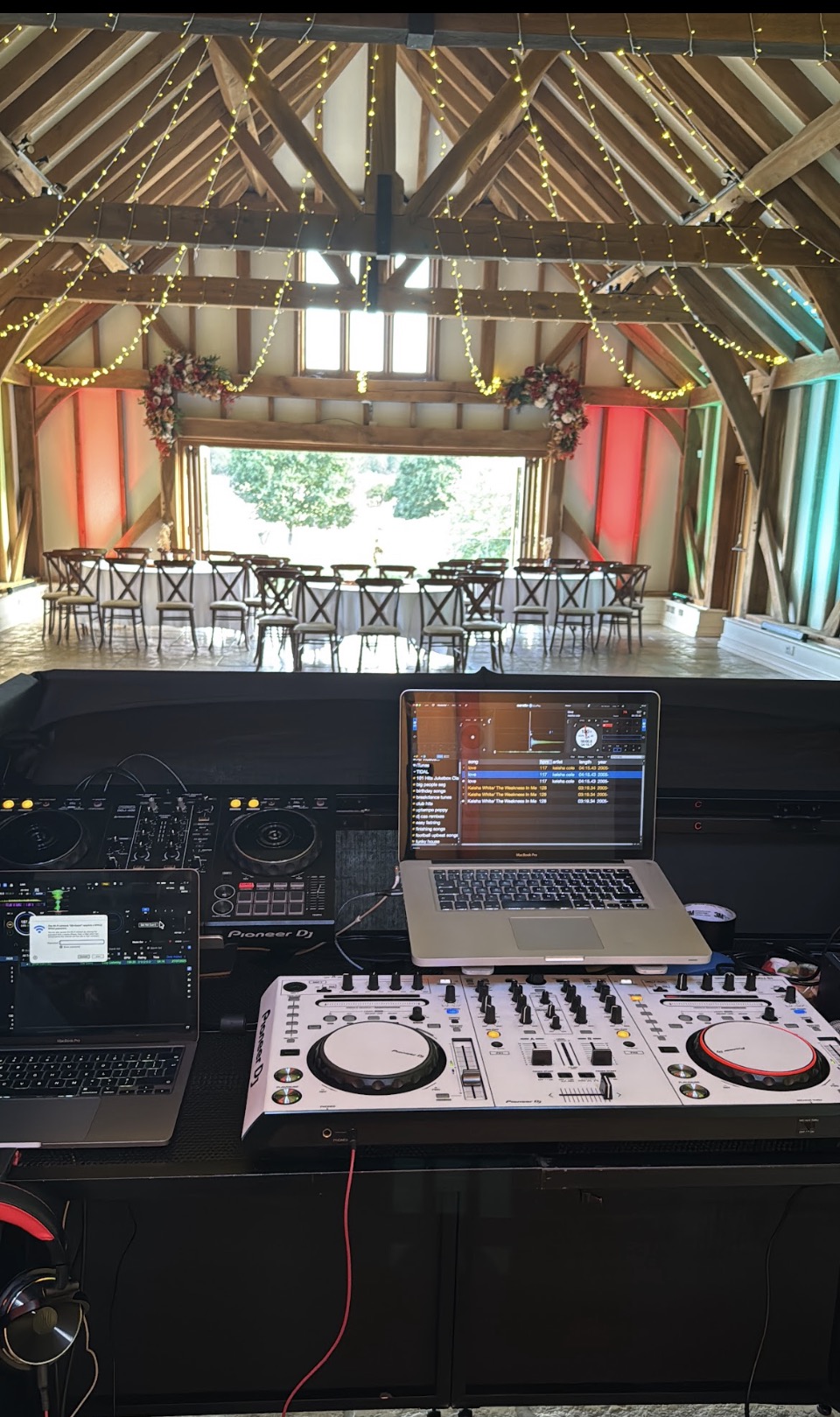 DJ booth setup at dusk with dual speakers, laptop showing waveform, LED uplights and fog haze blurred in background, wide 3:2 composition, photorealistic style demonstrating scale, safety rigging, and festival-grade production.