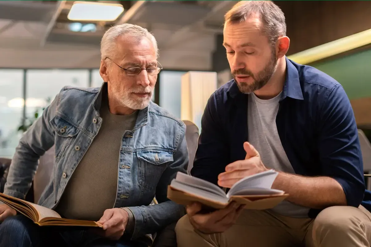 Two men sitting together indoors, reading and discussing a book.