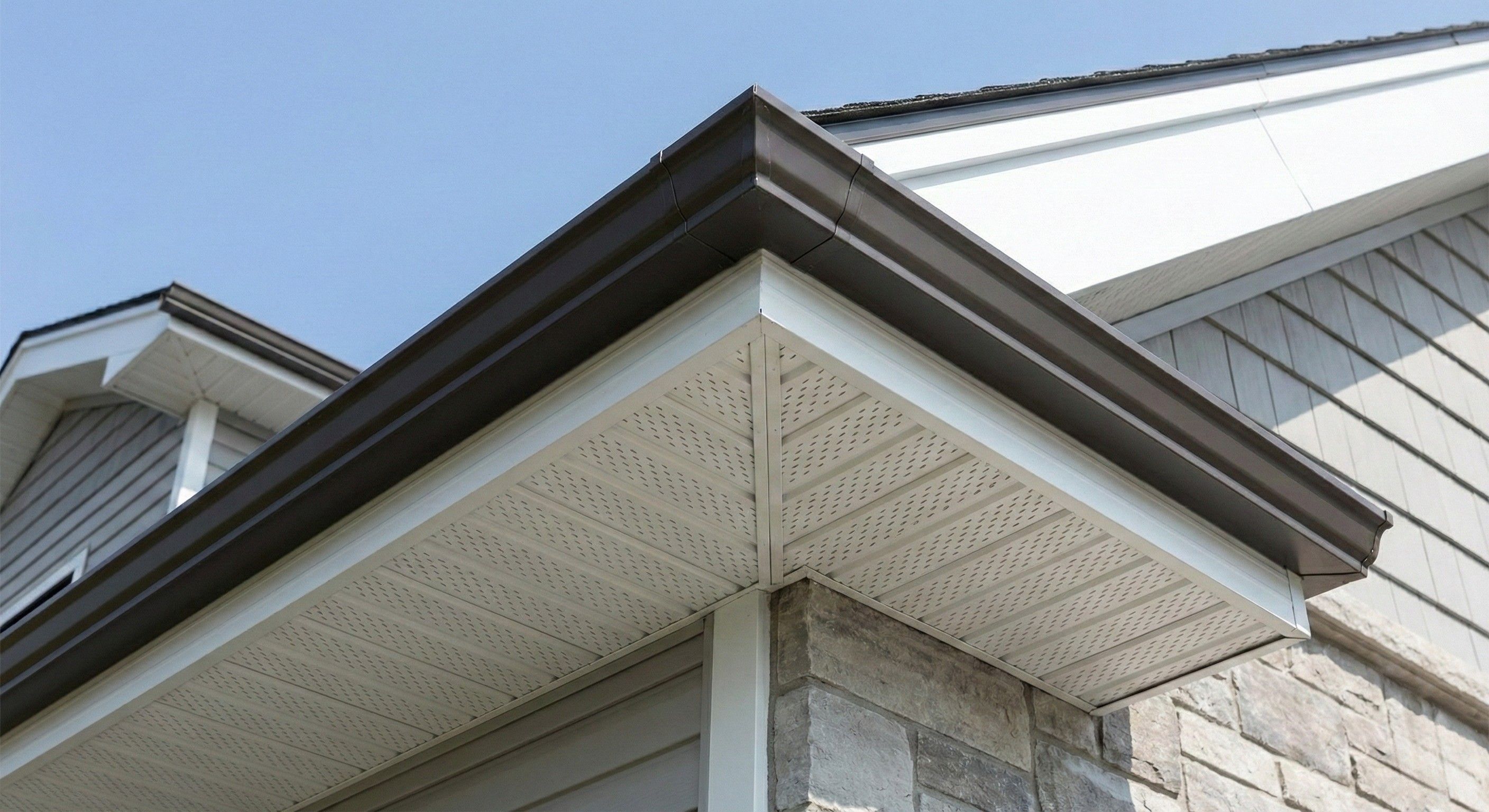 Close-up photograph of a newly installed dark bronze seamless aluminum eavestrough and white vented soffit system on a modern home with stone veneer by Ollie's Roofing in Milton.