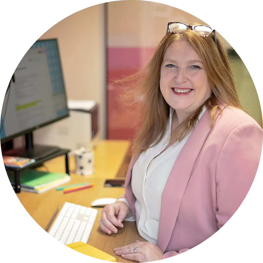 A woman with long red hair, smiling and seated at a desk, wearing a light pink blazer and a white blouse. She has glasses resting on her head and is surrounded by office supplies such as a keyboard, pens, a notebook, and a cup. In the background, there’s a computer screen showing a webpage and a soft-colored office setting.