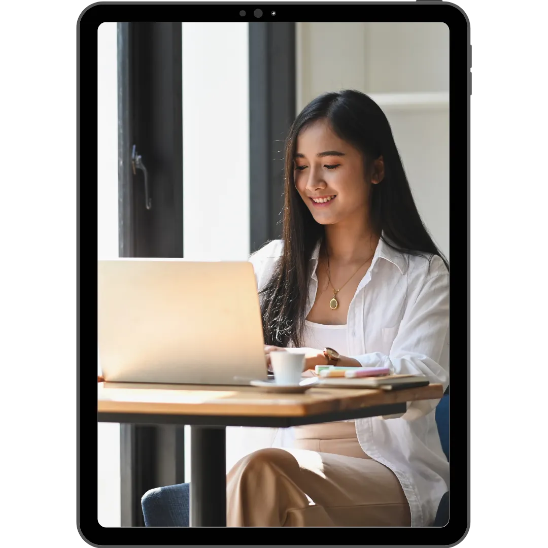 A woman with long black hair, sitting at a desk and smiling while working on a laptop. She is wearing a white shirt, a pendant necklace, and beige pants. A cup of coffee is placed on the desk in front of her, with stationery items like pens and a notebook nearby. Sunlight is streaming through the windows in the background, giving the image a warm, relaxed atmosphere.