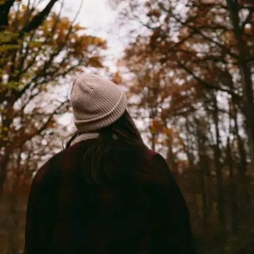 A person wearing a knit hat and dark jacket stands in a forest with autumn-colored trees, facing away from the camera.