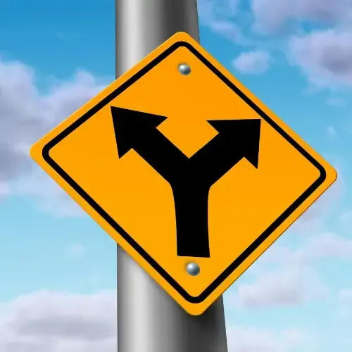 A yellow road sign with a black Y-shaped arrow, mounted on a pole against a blue sky with scattered clouds.