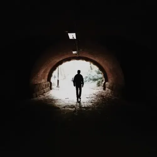 A person walking through a dark tunnel toward a bright light at the tunnel’s exit, with the tunnel entrance framed by stone.