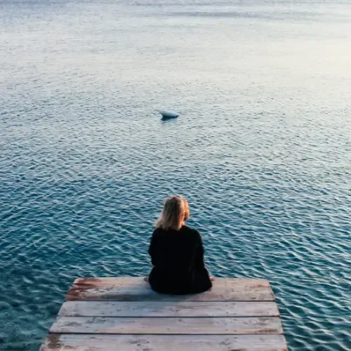 A person with blonde hair sits alone on the edge of a wooden dock, facing a calm, expansive body of water.