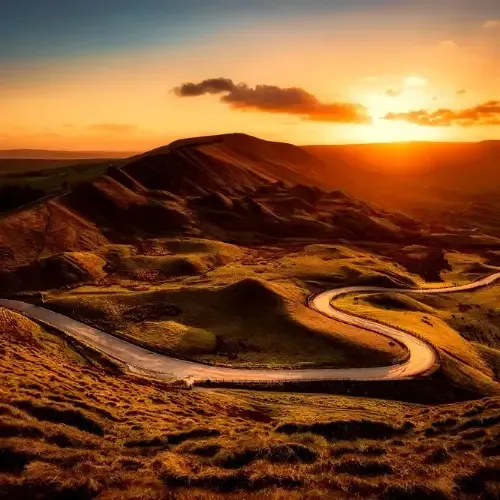 A winding road curves through a hilly landscape at sunset, with warm sunlight casting long shadows over the terrain.