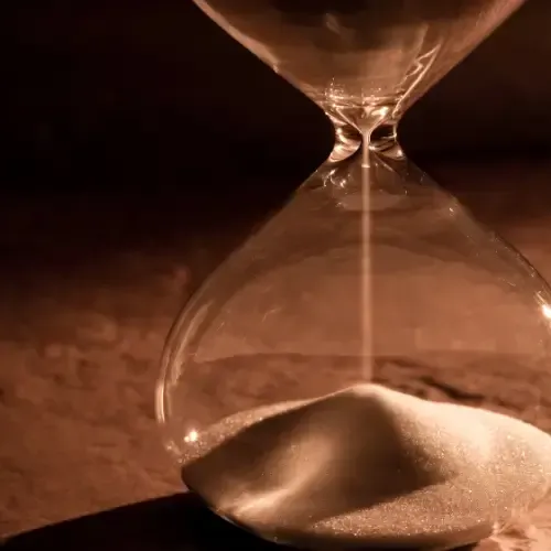 An hourglass with sand flowing from the top bulb to the bottom, placed on a dark surface with warm lighting.