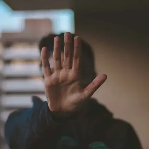 A person in a dark hoodie holding up an open hand toward the camera, obscuring their face, with a blurred urban background.
