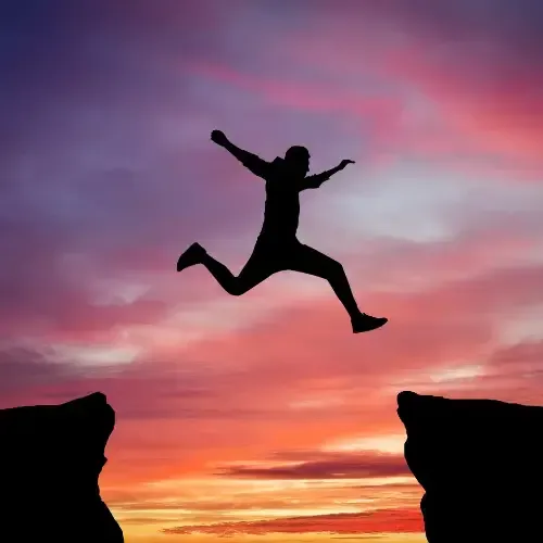 Silhouette of a person mid-air, leaping between two cliffs at sunset with a vibrant sky in the background.