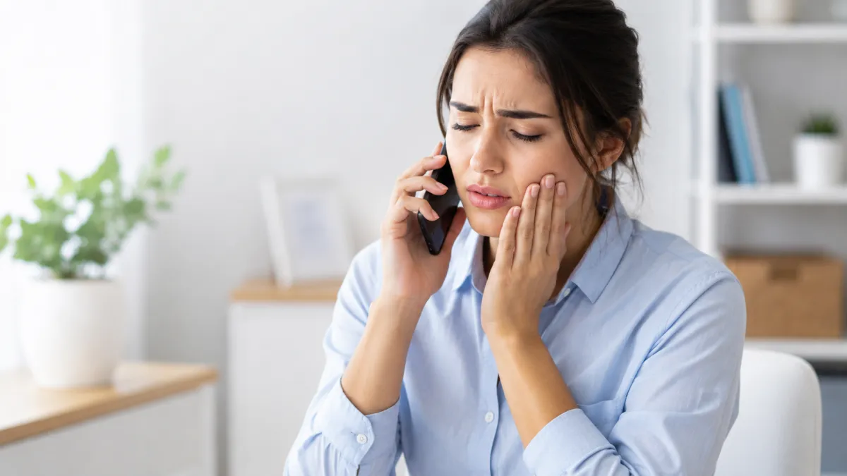 A person calling a dentist while holding their cheek, indicating a dental emergency