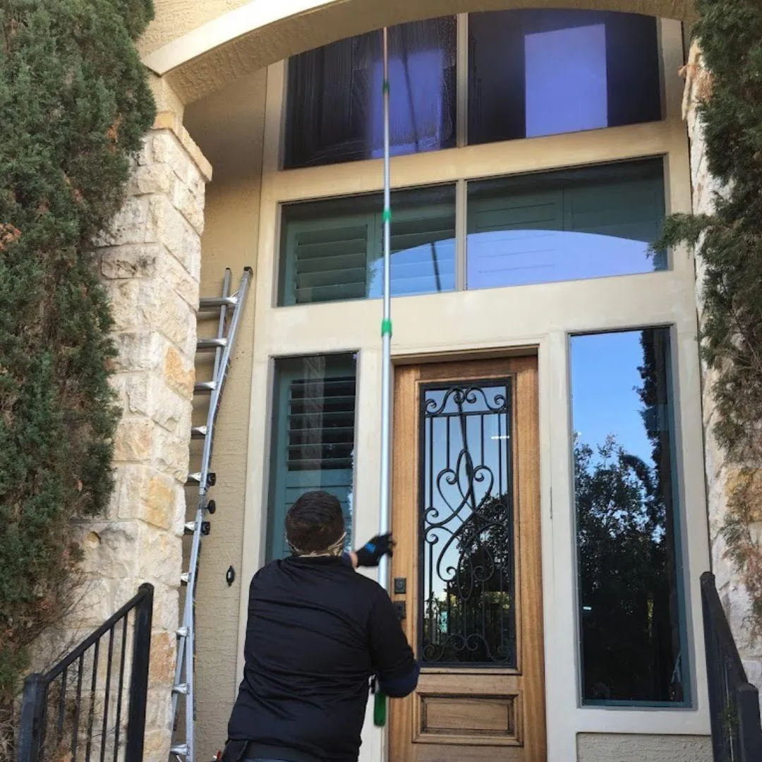 Professional window cleaner using telescopic pole to clean multi-story residential windows in San Antonio with specialized equipment" Option 2 (Service-focused): "D&D Window Cleaning technician performing exterior window cleaning service on luxury home in San Antonio using professional tools
