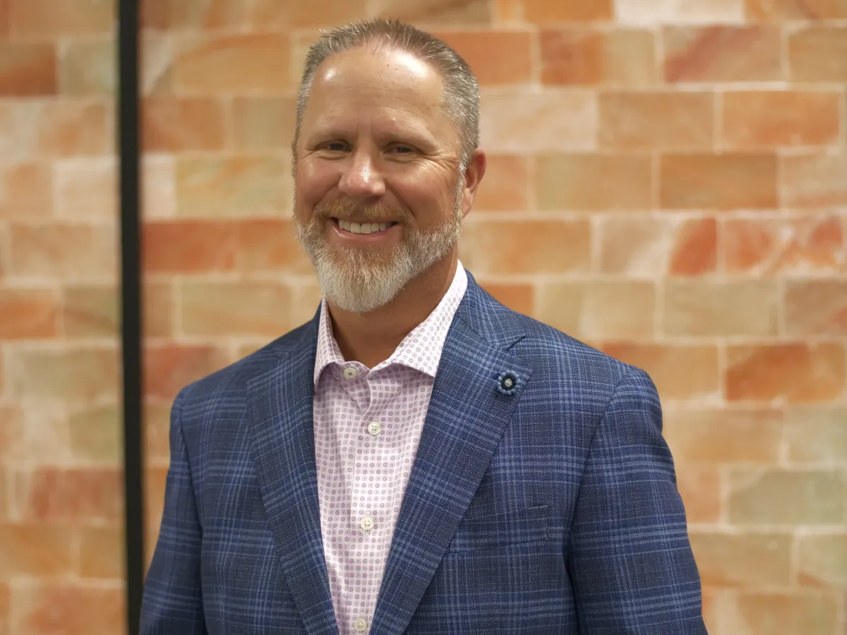 A smiling man with short gray hair and a beard, wearing a blue checkered suit jacket and light pink shirt, standing in front of a brick-patterned wall