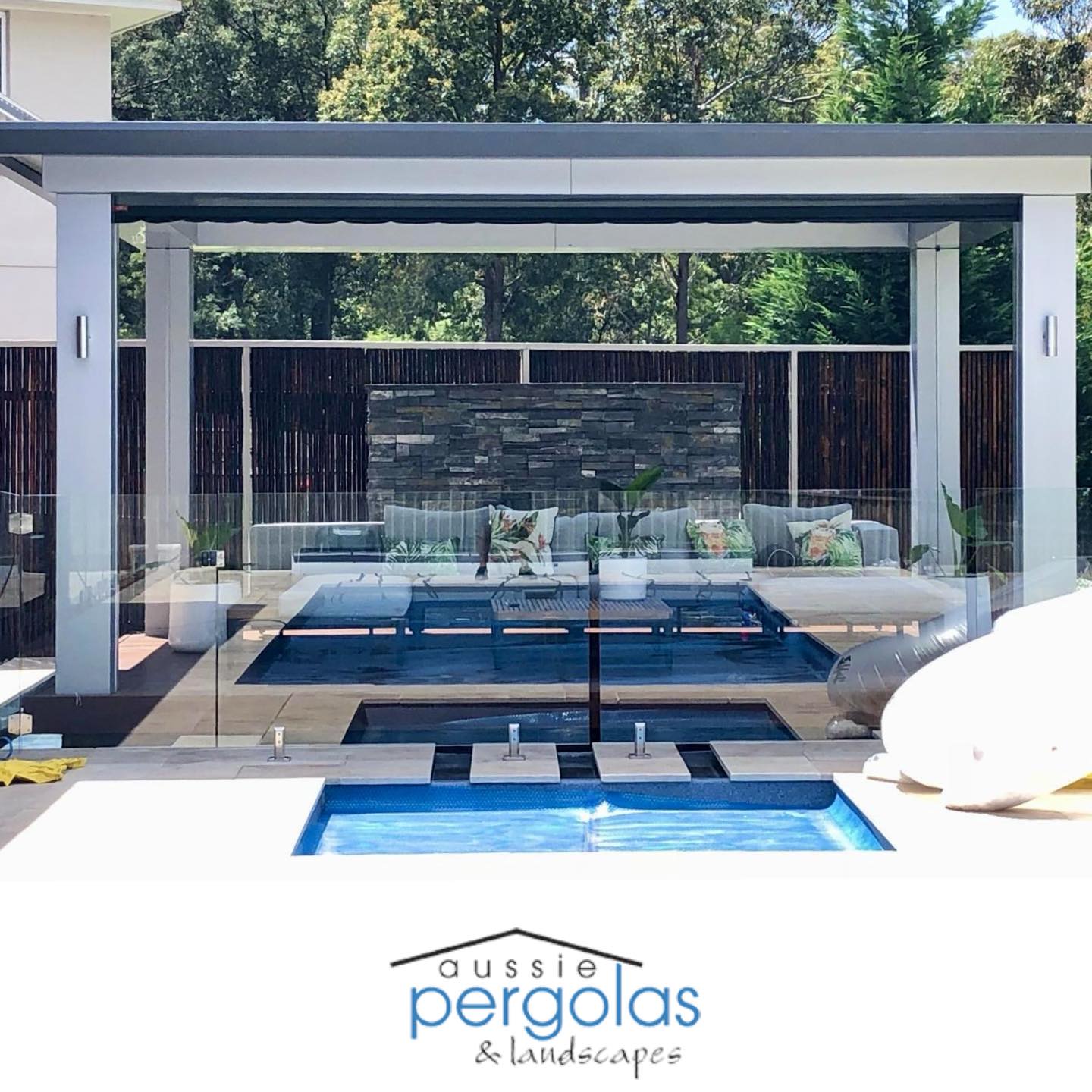 Modern pool pergola with stylish seating area, glass fencing, and landscaped backdrop, showcasing Aussie Pergolas branding.