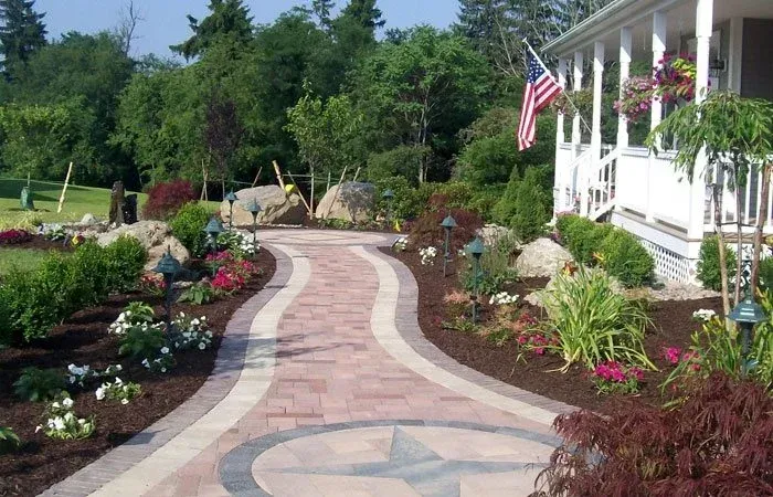 Curved stone pathway lined with colorful flower beds and greenery, leading to a porch with American flag, showcasing professional landscaping and garden design.