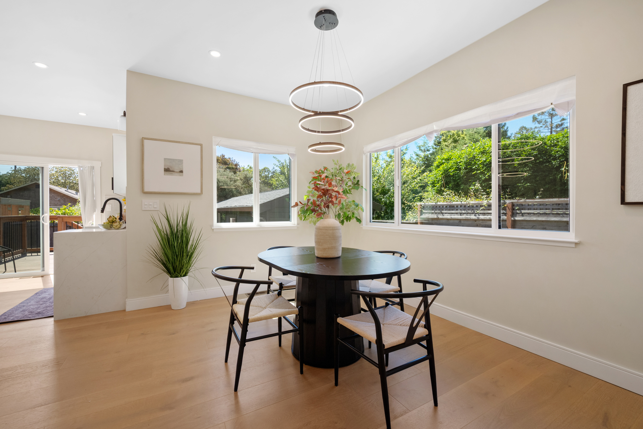 Modern dining area with large windows, ring chandelier lighting, wood flooring, and open kitchen connection by Casa de RJ Construction
