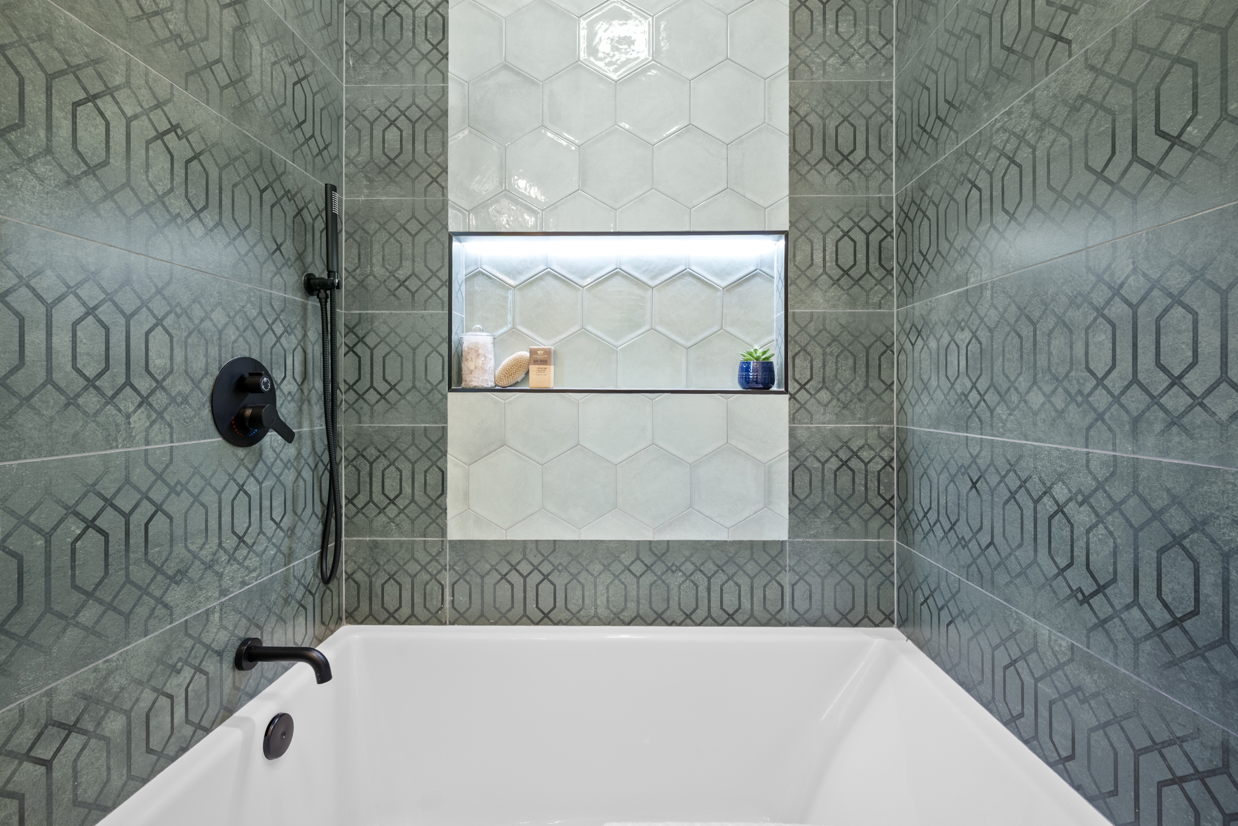 Modern bathroom tub shower with patterned tile walls, hexagon tile niche with lighting, and matte black fixtures by Casa de RJ Construction