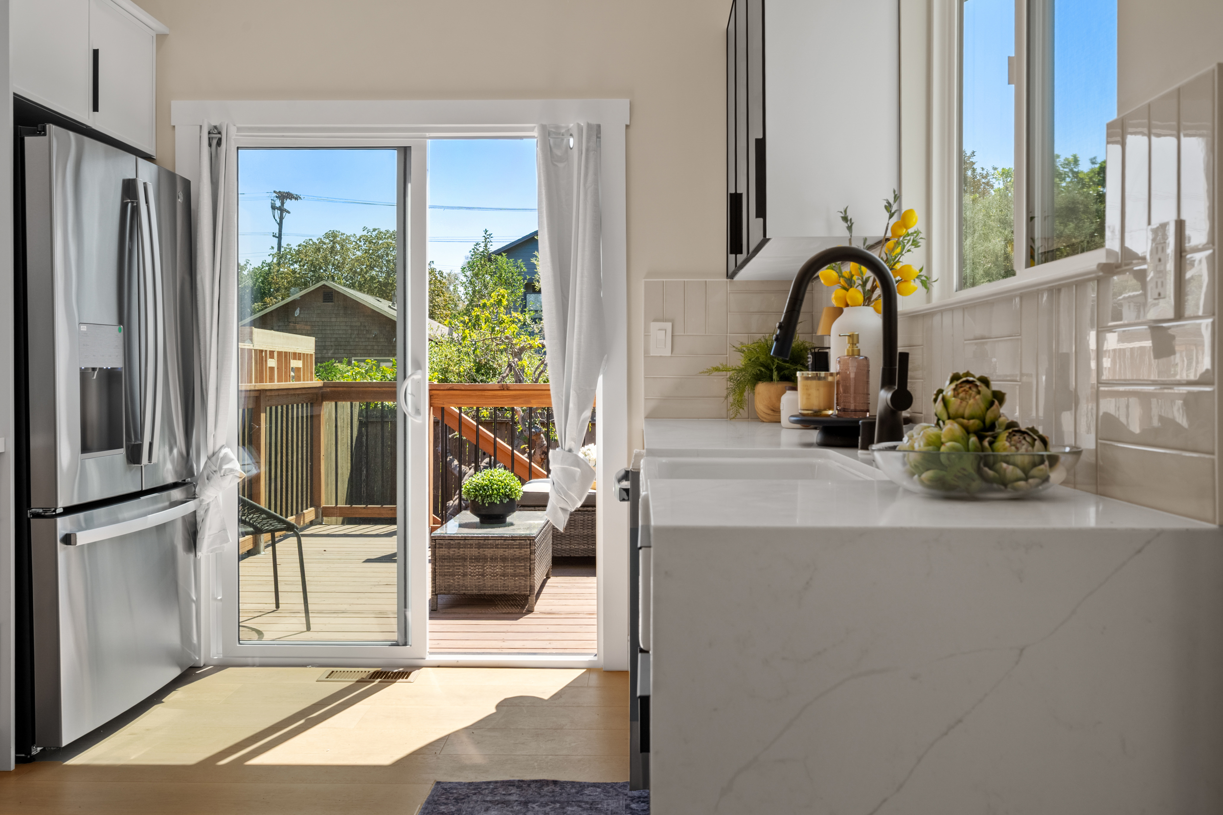 Modern kitchen with quartz counters, black faucet, white cabinets, stainless steel fridge, and sliding glass door to outdoor deck by Casa de RJ Construction