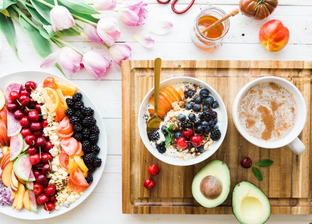 A colorful fruit plate next to a cutting board that has a white bowl with oatmeal with berries on top, a sliced open avocado, and a coffee drink with plant-based milk and cinnamon on top.