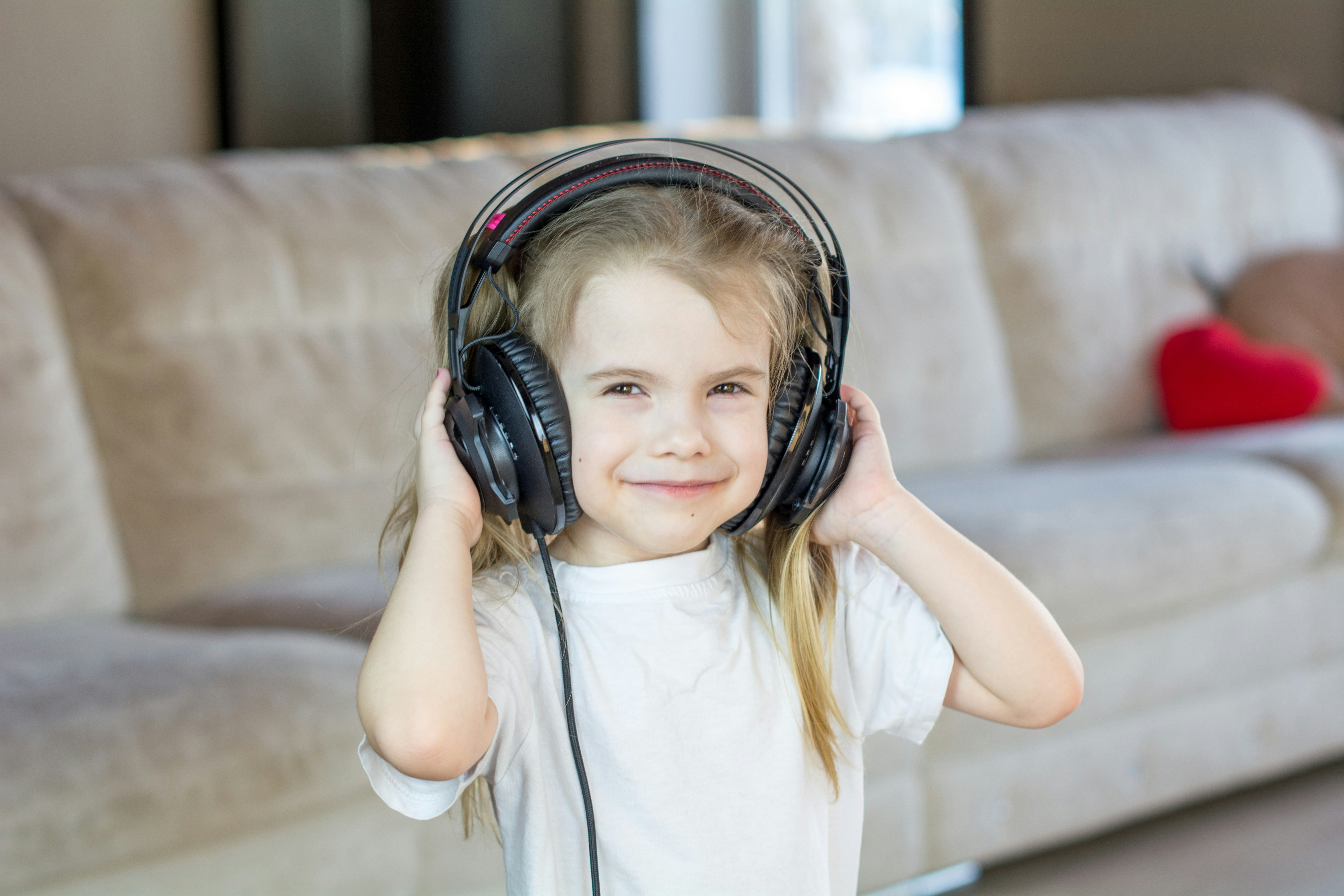 A preschool girl smiling while listening to music on large headphones at home.