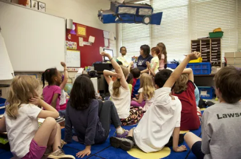 Preschool children sitting in a classroom circle, raising their hands while listening to their teacher