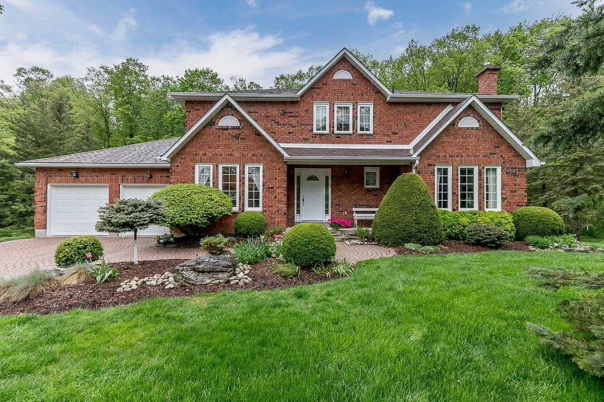 2-storey red brick detached home with attached 2-car garage, mature gardens, and paved brick driveway and walkway in the Purple Hill neighbourhood of Mono 