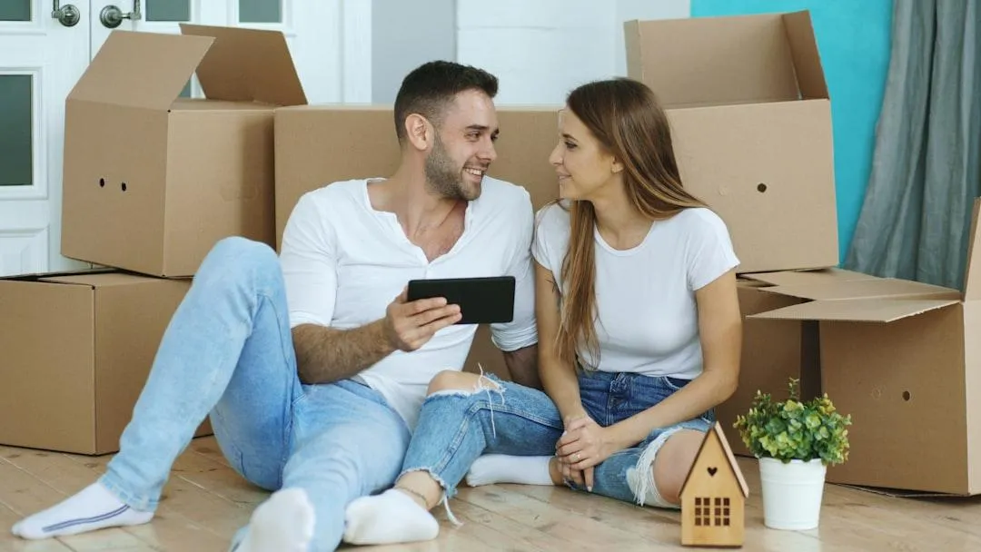 Couple sitting on the floor, looking up houses on a phone with moving boxes around them