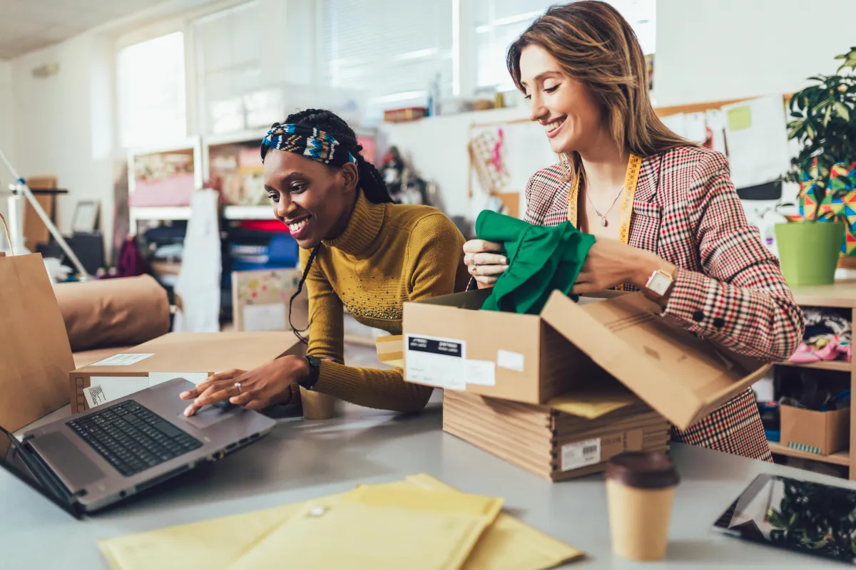 Black lady working on computer while white lady puts items into a shipping box
