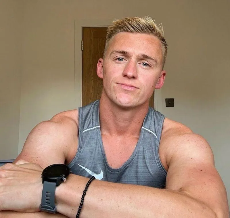 George Eastwood, a young white man with blonde hair. He is physically fit, wearing a sports shirt and fitness watch. 